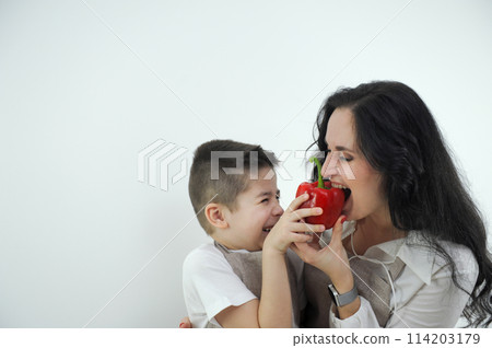 two friends mother biting bell pepper boy grimacing hugging space for text fun spending time together joke grocery store delicious vegetables vegetarian food wearing apron in kitchen mother and son 114203179