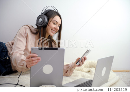 Smiling female student wears wireless headphones to study online with a Skype teacher, happy young woman learns a language, listen to a lecture watch a webinar, write notes, look at a laptop Smiling female student wears wireless headphones to study online with a Skype teacher, happy young woman learns a language, listen to a lecture watch a webinar, write notes, look at a laptop 114203229