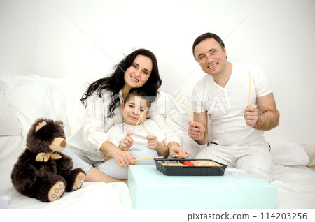home delivery of sushi happy cheerful family eating sushi from disposable tableware with chopsticks toy teddy bear nearby european family happy eating in bed day off spending time with family idyll 114203256