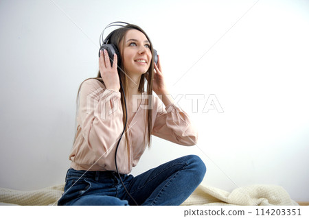 a girl in blue jeans sits on the floor on blanket on the bed raised her head up listens to music on headphones place for text online work conference attractive girl with headphones on gray background. 114203351