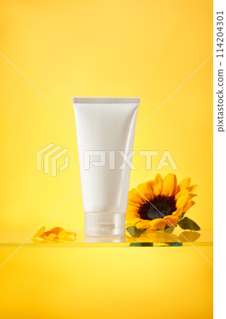Unlabeled white bottle displayed on glass surface isolated over yellow background, decorated by a fresh sunflower and some sunflower petals. Mockup for advertising with front view 114204301
