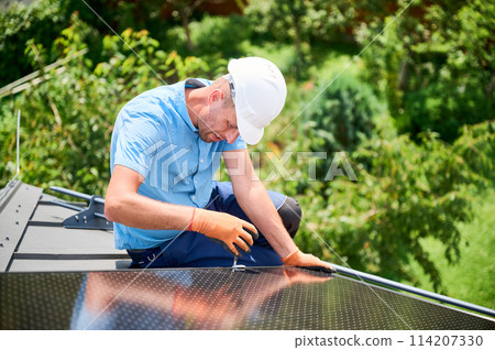 Worker building photovoltaic solar panel system on rooftop of house. Man engineer in helmets and gloves installing solar module with help of hex key outdoors. Alternative energy generation concept. Worker building photovoltaic solar panel system on rooftop of house. Man engineer in helmets and gloves installing solar module with help of hex key outdoors. Alternative energy generation concept. 114207330