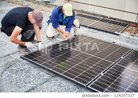 Workers building photovoltaic solar panel system on metal rooftop of house. Men engineers installing solar module with help of wrench outdoors. Alternative and renewable energy generation concept. Workers building photovoltaic solar panel system on metal rooftop of house. Men engineers installing solar module with help of wrench outdoors. Alternative and renewable energy generation concept. 114207337