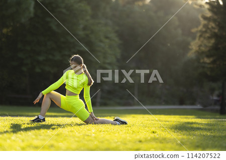 A woman in neon yellow is stretching her legs on the grass in the park A woman in neon yellow is stretching her legs on the grass in the park 114207522