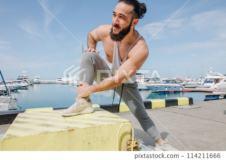 Sports lifestyle. Happy young caucasian bodybuilder stretching on the sea shore. Sports lifestyle. Happy young caucasian bodybuilder stretching on the sea shore. 114211666