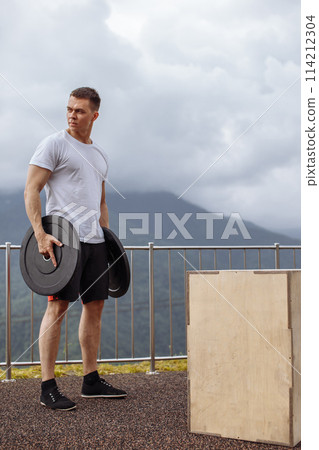 Strong male athlete doing box jumps with two plates outdoor on top of mountain. Strong male athlete doing box jumps with two plates outdoor on top of mountain. 114212304