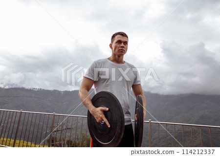 Stong male athlete pumping musculs with weight plates in outdoor workout. 114212337