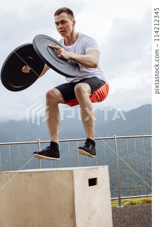 Strong male athlete doing box jumps with two plates outdoor on top of mountain. Strong male athlete doing box jumps with two plates outdoor on top of mountain. 114212345