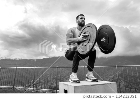 Strong male athlete doing box jumps with two plates outdoor on top of mountain. 114212351