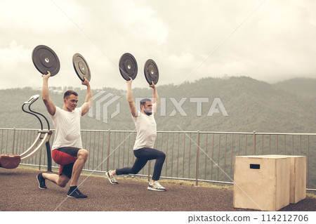 two athletes with barbell disc making lunges in outdoor workout 114212706