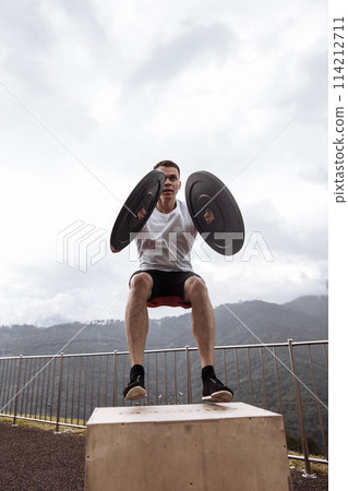 Strong male athlete doing box jumps with two plates outdoor on top of mountain. 114212711