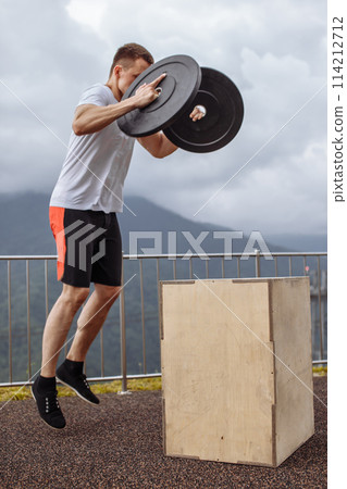 Strong male athlete doing box jumps with two plates outdoor on top of mountain. 114212712