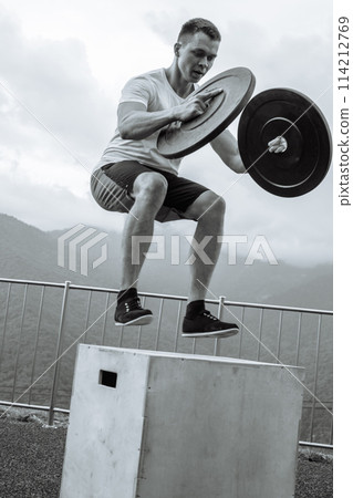 Strong male athlete doing box jumps with two plates outdoor on top of mountain. 114212769