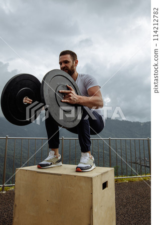 Strong male athlete doing box jumps with two plates outdoor on top of mountain. 114212782