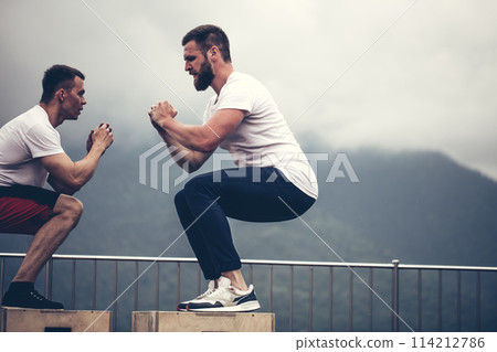 Two male athletic friends doing box jump exercise outdoor, in mountains 114212786