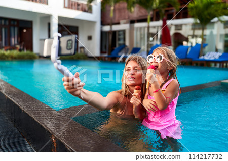 Blogger mom snaps selfie with daughter eating ice cream in pool at luxury resort. Smiling duo shares summer moment for social media. Casual holiday fun with family, leisure in tropical paradise. 114217732