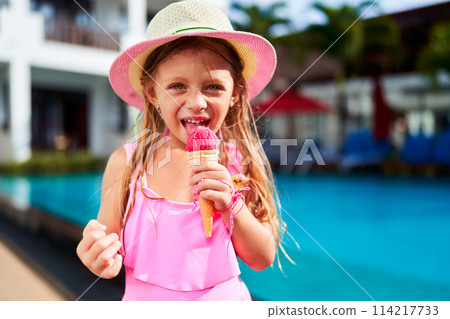 Little girl in pink swimsuit enjoys strawberry ice cream by poolside. Happy child relaxing after swim, savoring sweet treat on sunny day. Summer vacation, outdoor leisure for kids, refreshment. Little girl in pink swimsuit enjoys strawberry ice cream by poolside. Happy child relaxing after swim, savoring sweet treat on sunny day. Summer vacation, outdoor leisure for kids, refreshment. 114217733