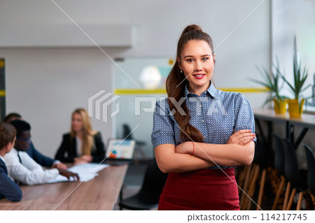 Confident Asian businesswoman stands in modern office, arms crossed. Teamwork in background, diverse colleagues discuss project. Professional meeting in corporate environment, leadership, startup. Confident Asian businesswoman stands in modern office, arms crossed. Teamwork in background, diverse colleagues discuss project. Professional meeting in corporate environment, leadership, startup. 114217745