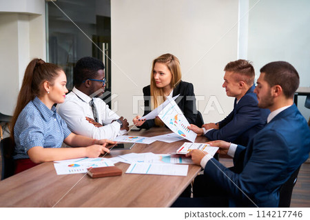 Multiracial team analyzes graphs in office, African man in suit shares report with diverse colleagues. Mixed group engages in strategy discussion, women and men in corporate attire collaborate Multiracial team analyzes graphs in office, African man in suit shares report with diverse colleagues. Mixed group engages in strategy discussion, women and men in corporate attire collaborate 114217746