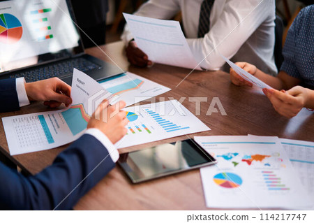 Multiethnic team analyzes business metrics at workplace, discussing colorful graphs on reports, planning strategy. Corporate group collaborates, sharing insights, focusing on data-driven solutions. 114217747