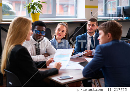 Multiethnic corporate team in meeting room discuss strategy. Diverse group of businesspeople with charts engage in brainstorming, collaboration. A focused team analyzes data for project planning. 114217755