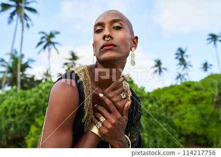 Trans sexual ethnic fashion model with brass jewelry accessories in elegant posture looks at camera. Epatage gay black man with golden rings, earrings, bracelets touches shoulder on scenic sea beach 114217756