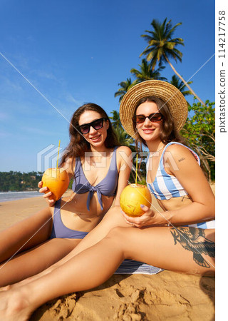 Two women in bikinis enjoy fresh coconuts on sunny tropical beach. Friends sit on golden sand under blue sky, embrace summer vibes. Travel lifestyle, relaxation with exotic drinks by sea. 114217758