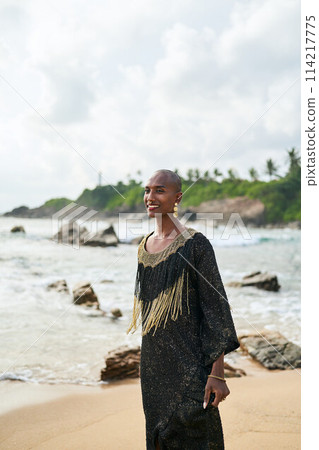 Flamboyant gay black male in luxury gown poses on scenic ocean beach. Trans sexual ethnic fashion model in long posh dress and accessories looks at a camera. Sea waves and lighthouse on background. Flamboyant gay black male in luxury gown poses on scenic ocean beach. Trans sexual ethnic fashion model in long posh dress and accessories looks at a camera. Sea waves and lighthouse on background. 114217775