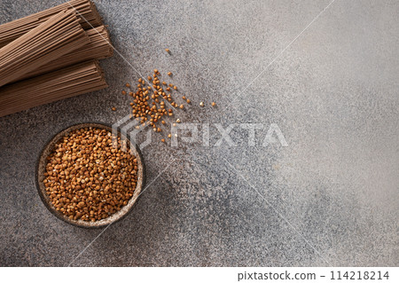Uncooked buckwheat noodles, soba and grain in bowl on dark gray background. View from above. Space for text. Healthy vegan diet. Alternative gluten free pasta. Uncooked buckwheat noodles, soba and grain in bowl on dark gray background. View from above. Space for text. Healthy vegan diet. Alternative gluten free pasta. 114218214