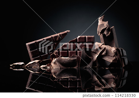 A broken chocolate bar and pieces of dark chocolate on a black reflective background. A broken chocolate bar and pieces of dark chocolate on a black reflective background. 114218330