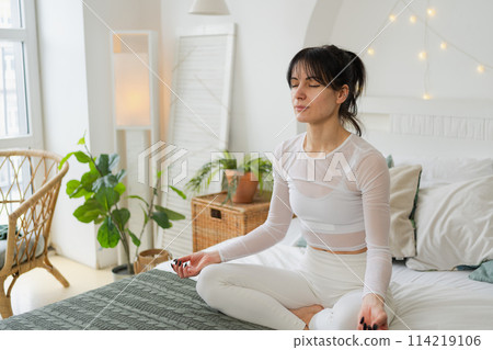 Yoga mindfulness meditation. Young healthy woman practicing yoga in bedroom at home. Woman sitting in lotus pose on bed meditating smiling relaxing indoor. Girl doing breathing practice. Yoga at home Yoga mindfulness meditation. Young healthy woman practicing yoga in bedroom at home. Woman sitting in lotus pose on bed meditating smiling relaxing indoor. Girl doing breathing practice. Yoga at home 114219106