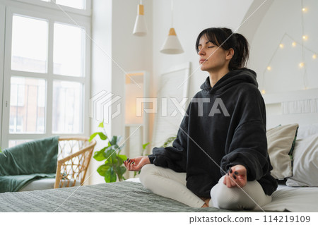 Yoga mindfulness meditation. Young healthy woman practicing yoga in bedroom at home. Woman sitting in lotus pose on bed meditating smiling relaxing indoor. Girl doing breathing practice. Yoga at home 114219109