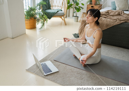 Yoga mindfulness meditation online. Woman practicing yoga with online lessons in laptop at home. Woman sitting in lotus pose meditating relaxing indoor. Girl doing breathing practice. Yoga at home 114219111