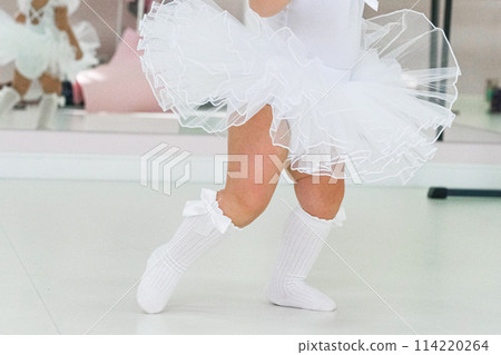 Portrait of a cute baby ballerina sitting near the ballet barre putting on shoes 114220264