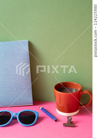 Desktop workspace. notebook, eyeglasses, colored pencil, clip, cup of hot chocolate on pink desk. khaki green wall background 114225980
