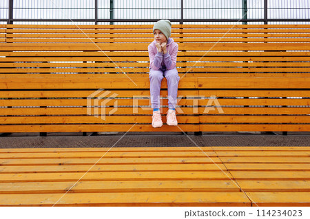 A girl sits on a bench in a sports tribune with her hands on her knees. 114234023