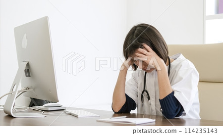 Stressed female doctor suffers from headache sitting at desk in clinic 114235143