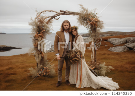 Boho style bride and groom are holding hands in the background of romantic wedding arch at the sea bay and mountains. 114240350
