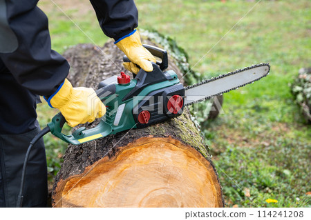 A man in uniform cuts an old tree in the yard with an electric saw 114241208