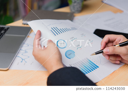 Close-up of a professional hand reviewing business document data performance analytics and printing statistical reports and planning marketing and financial strategy on a desk. Close-up of a professional hand reviewing business document data performance analytics and printing statistical reports and planning marketing and financial strategy on a desk. 114243030