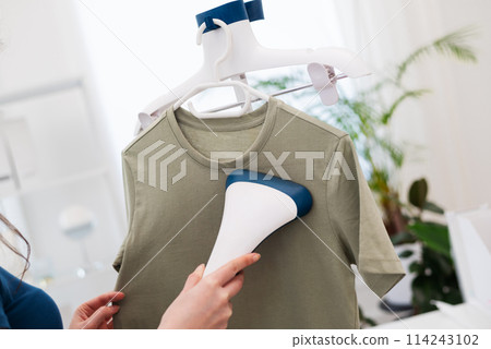 Cropped woman using steaming iron to ironing fashion shirt at home in bright cozy room. Female holding electric steamer Cropped woman using steaming iron to ironing fashion shirt at home in bright cozy room. Female holding electric steamer 114243102