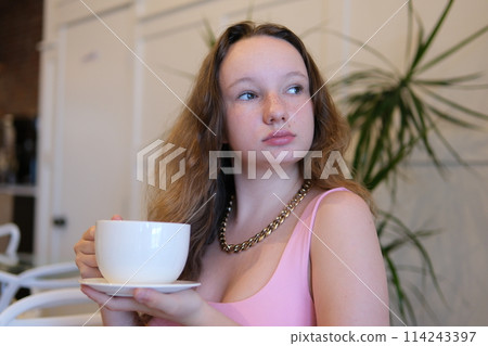 A young girl in a pink dress enjoying cappuccino in a street cafe drinks tea from a large mug of cappuccino latte in a restaurant. European young woman drinks chocolate milk in a cafe. High quality 114243397