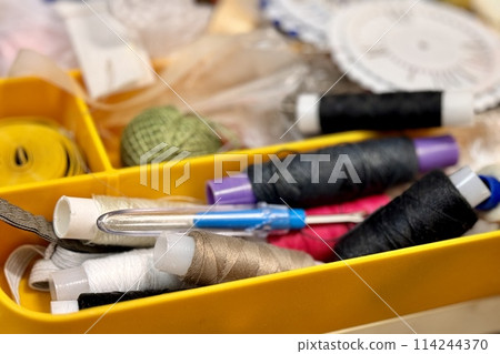 A lot of sewing items such as buttons, colorful fabrics, scissors, measuring tape, thimble, spools of thread on sewing pattern located in plastic box on the sofa. Close-up 114244370