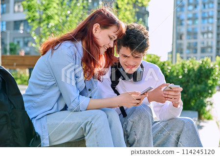 Teenage youth guy and girl college students sitting outdoor using smartphone Teenage youth guy and girl college students sitting outdoor using smartphone 114245120