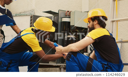 Skilled expert assembling professional pressure benchmarking tool to check liquids and gases in hvac system while african american mechanic cleans layer of built up dirt and dust in condenser 114246029