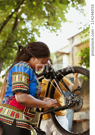 Black woman outdoors checking and fixing bicycle wheel with professional equipments for summer cycling. Young african american woman repairing broken bike tire and chains with tools in yard. Black woman outdoors checking and fixing bicycle wheel with professional equipments for summer cycling. Young african american woman repairing broken bike tire and chains with tools in yard. 114246058