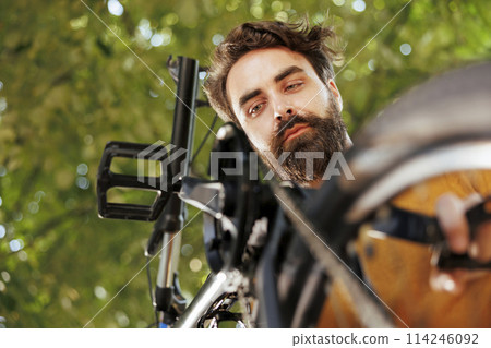 Active caucasian male working on broken bike wheel chain ring with professional equipment in home yard. Committed sporty man servicing bicycle outside for summer leisure cycling. 114246092