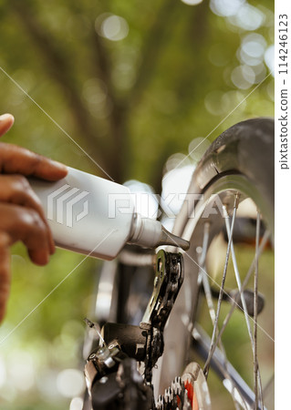 Detailed shot of bicycle chain ring being lubricated with specialized grease for safe outdoor leisure cycling. Image showing close-up view of african american hand holding chain lubricant. 114246123