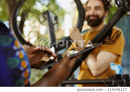 Closeup of black woman grasping phone tablet while enthusiastic young man in background repairs bicycle wheel outdoors. African american woman holding digital device to research fixes for broken bike 114246127