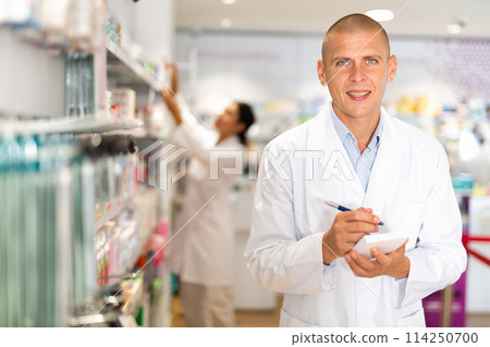 Portrait of male pharmacist working in pharmacy during the pandemic, standing in trading floor and makes important notes 114250700